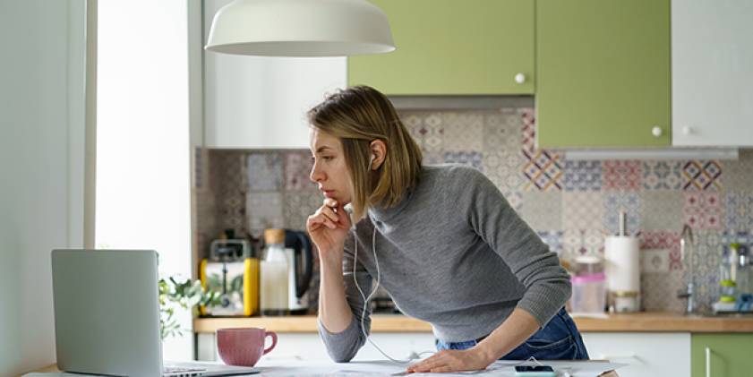 Menyw gyda chlustffonau yn syllu ar liniadur yn y gegin