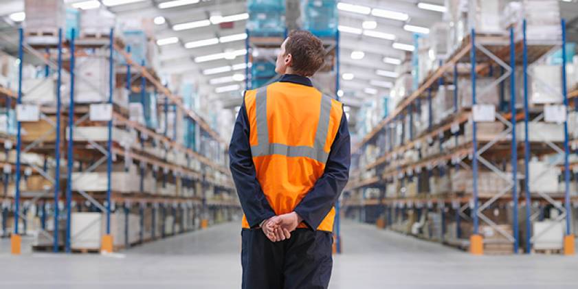 Gweithiwr warws mewn tiwnig oren yn wynebu eiliau o silffoedd.